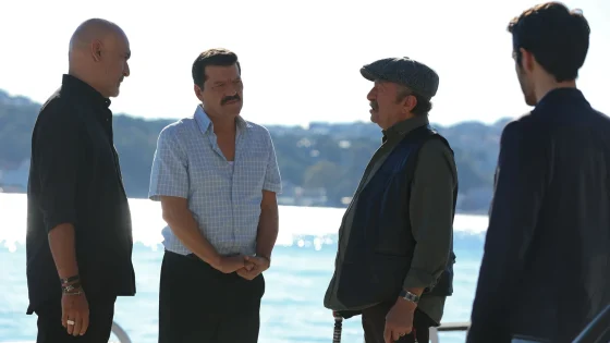 Four men stand in a circle by the water, engaged in conversation outdoors on a sunny day.