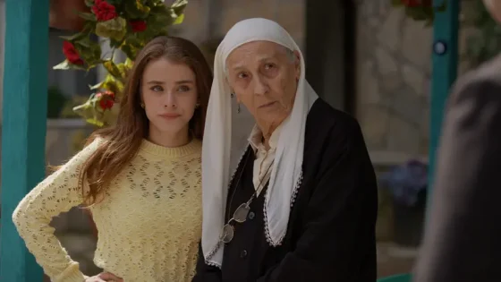 Young woman in a yellow lace sweater stands with an elderly nun in a black outfit and white headscarf, outdoors and looking concerned.