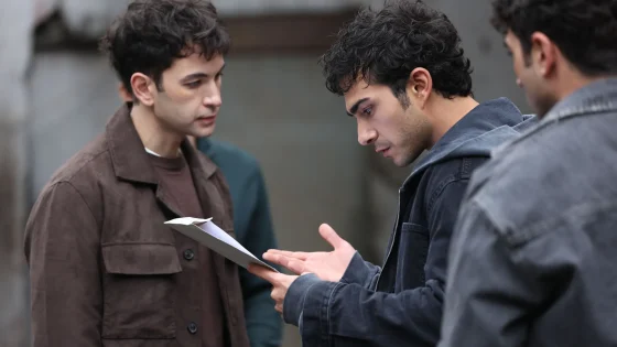 Three men converse outdoors, one holds a paper while another gestures with an open hand.