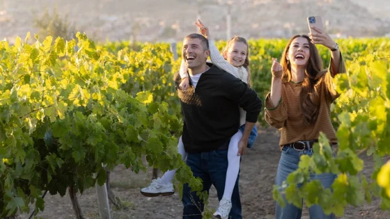 Family of three taking a selfie in a vineyard: father carrying daughter on his back while mother snaps the photo with her phone, all smiling to the camera.