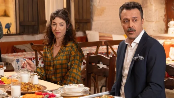 A woman and a man sit at a dining table in a warmly lit restaurant, with plates of food and glasses in front of them, looking slightly to the side.