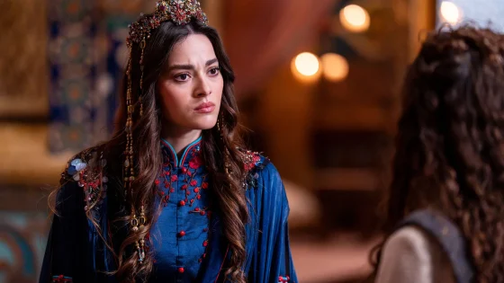 Woman wearing an ornate jeweled crown and blue embroidered traditional dress, facing left with a thoughtful expression, warm bokeh lights in the background
