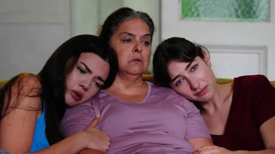 Three women sit on a couch, leaning on each other with somber expressions; the middle woman wears a lavender shirt.