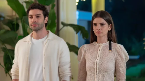 Two adults stand side by side in a well-lit room, looking serious as if in a moment of conversation or tension.