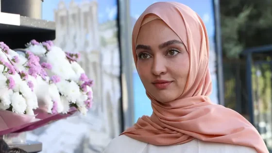 Portrait of a woman wearing a peach hijab, standing beside a bouquet of white and pink flowers outdoors.