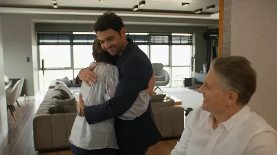 Two people share a warm hug in a bright, modern living space while a smiling man watches in the foreground, creating a cozy scene.