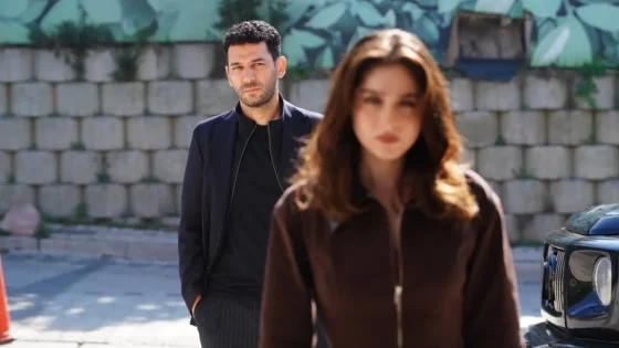 Man in a dark blazer stands behind a blurred woman in a brown jacket near a parked blue car, outdoors by a stone wall.