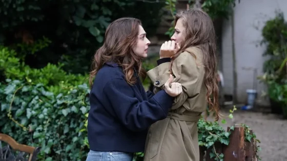 Two women confront each other in a garden, one grabbing the other's coat sleeves in a tense moment outdoors.