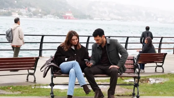 A couple sits on a park bench by the water, looking at a phone together with a foggy shoreline in the background.