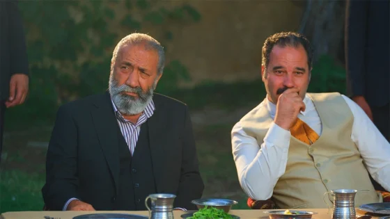 Two men sit outdoors at a table, one with a gray beard in a dark suit and the other in a beige vest and white shirt, with metal cups on the table.