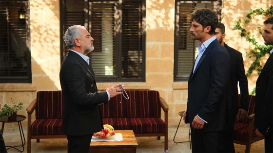 Older man in a dark suit holds prayer beads toward a younger man in a navy suit in a stone courtyard.