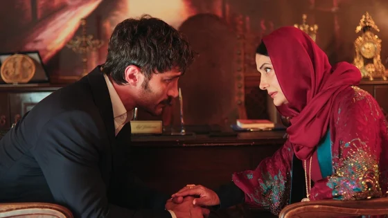 Man in a dark suit and a woman in a red headscarf sit facing each other, hands clasped in a respectful handshake in a warm, ornate room.