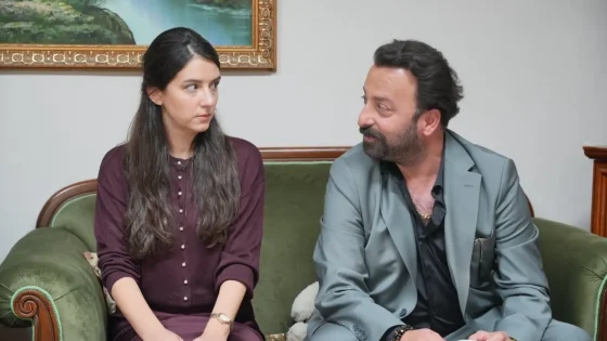 Woman in a burgundy dress and a man in a gray suit sit on a green velvet sofa indoors, facing each other as they talk; a framed landscape painting hangs behind them.