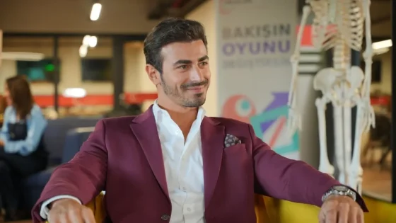 Man in a burgundy blazer sits relaxed on a yellow chair in a modern workspace, smiling. Skeleton model visible in background.
