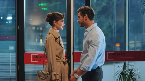 Two professionals face each other in a glass-door lobby: woman in a beige trench with a handbag and a man in a light blue shirt.