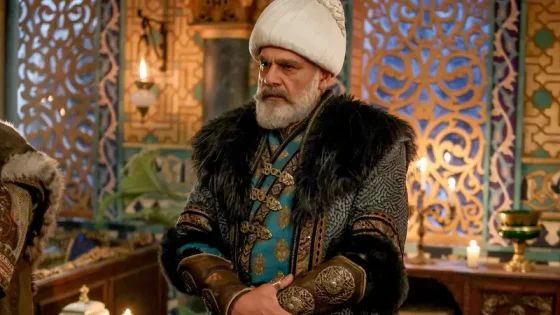 Older man with a white turban and gray beard in turquoise ceremonial robes and fur cloak, standing with folded hands in a richly decorated room.