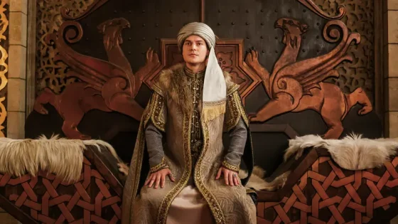 Man wearing a gold-embroidered robe and turban sits on a throne with carved dragon motifs behind him.