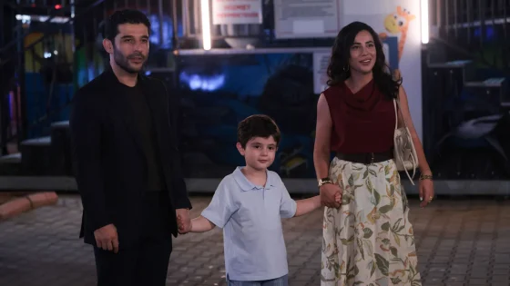 Family of three holding hands at night: man in a black jacket, woman in red sleeveless top and floral skirt, and a boy in light blue polo, smiling.