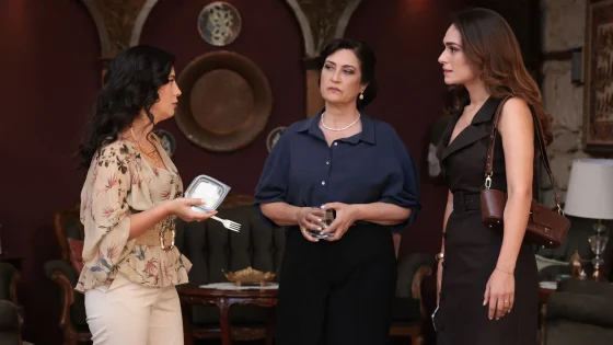 Three women converse at a social gathering: the left woman holds a takeout container and fork, the middle woman in a blue blouse watches, and the right woman wears a black sleeveless dress with a brown shoulder bag.