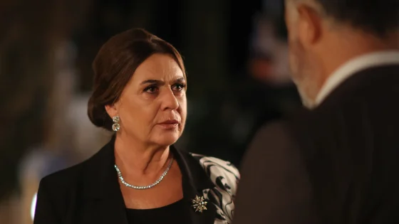 Close-up of a woman wearing diamond earrings and a necklace, looking up at a man in a suit at a dimly lit formal event.