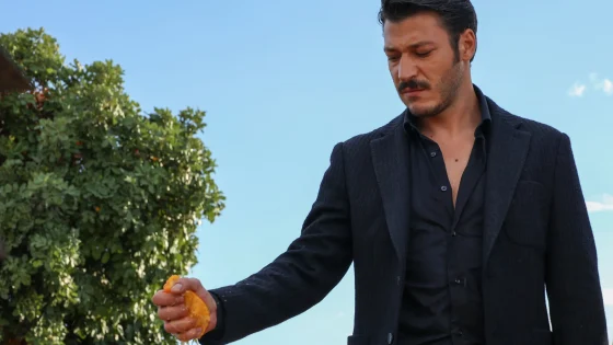 Man in a dark blazer holding an orange fruit outdoors with a clear blue sky and greenery to the left.