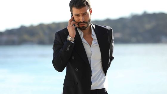Man in a dark suit talking on a smartphone by a sunny body of water, with a distant shoreline in the background.