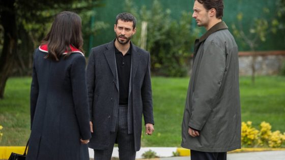 Three adults converse outdoors: a man in a dark coat center, a woman with her back to the camera, and a man in a gray coat to the right near a green lawn.