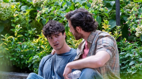 Two men sit among lush green plants as they converse, one with a hand on the other's shoulder.