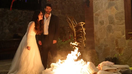 Bride in a white lace wedding dress and a man in a dark suit stand beside a bright indoor fire.