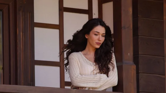 Woman with long dark hair in a cream knitted sweater stands beside a wooden building, looking down in a thoughtful pose.