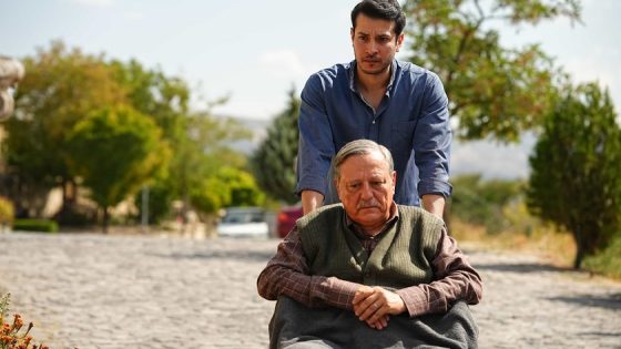 Younger man pushes an elderly man in a wheelchair along a sunlit cobblestone path outdoors, both looking ahead with calm expressions.