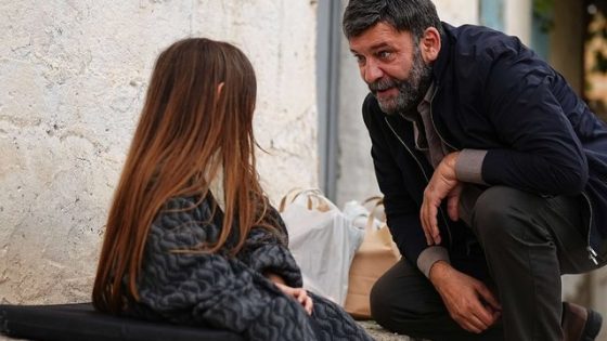 Man crouching to speak with a woman seated on a doorstep outside, near a rough wall and bags on the ground; a serious conversation.