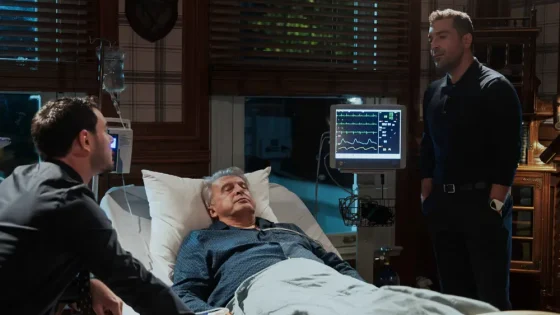 An elderly patient rests in a hospital bed while two visitors stand nearby and a monitor displays vital signs.
