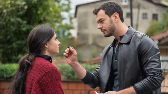 Man in a black leather jacket applies makeup or lipstick to a woman outdoors, near a brick wall and greenery behind them.