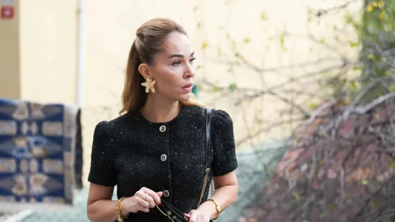 Stylish woman in a black textured dress stands outdoors, holding sunglasses and looking to the side.