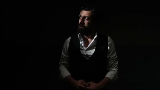 Bearded man in a white shirt and black vest sits in a dark setting, looking to the side with his hands clasped.