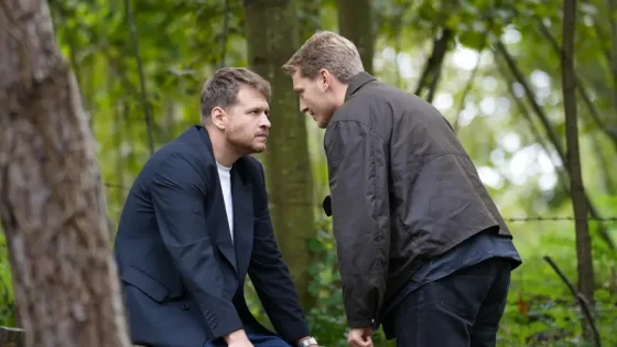 Two men lean toward each other in a tense confrontation in a wooded outdoor setting.