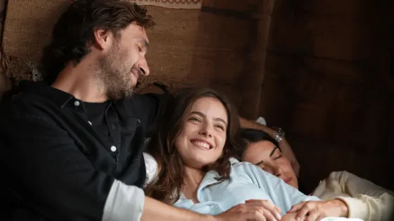 Three adults lounging on a couch, cuddled together and smiling warmly at the camera/each other, conveying a cozy, affectionate moment.