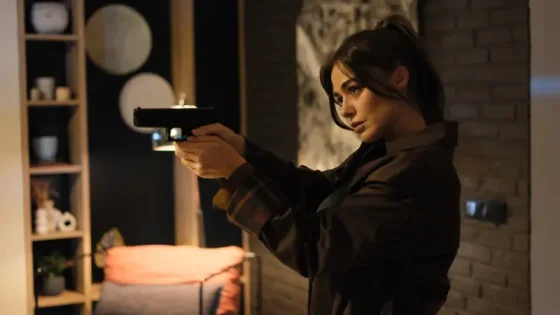 Woman indoors aiming a handgun with two hands, focused on target, in a dim living room setting.