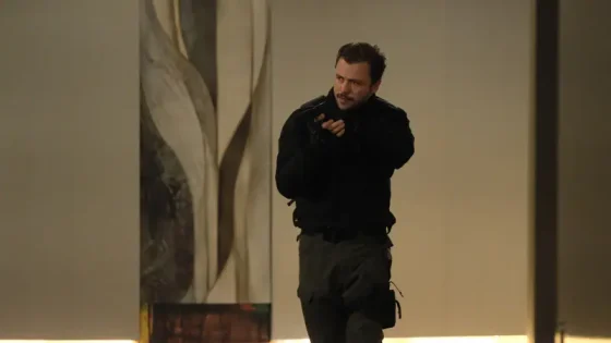 Man in tactical gear aims a handgun in a dim hallway, ready to fire, with a focused expression