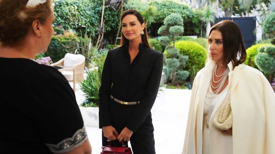 Two women in stylish outfits converse in a sunny garden while facing a third person seen from behind; one wears black, the other white with pearl necklaces and a red handbag visible.