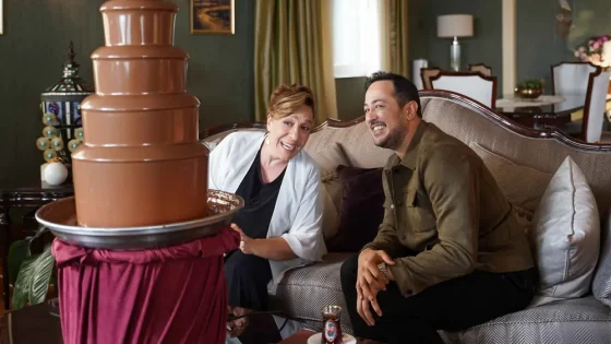 Smiling couple sits on a sofa beside a tall multi-tier chocolate fountain in a cozy living room. A Turkish tea glass rests on the coffee table nearby.