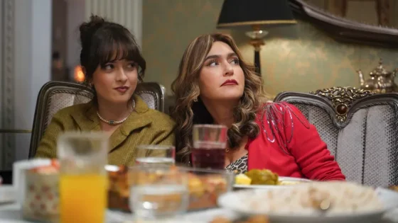 Two women sit at a fancy dining table with drinks and food, one in a mustard blazer and the other in a red jacket, looking to the side.