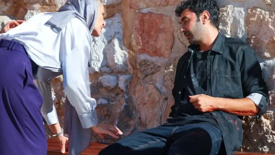 Woman wearing a light headscarf helps a man with a bleeding forearm who is sitting on a wooden bench against a stone wall outside.