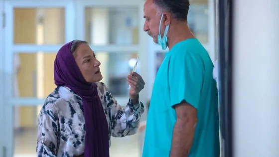 A woman in a purple headscarf offers a syringe to a male nurse in teal scrubs in a hospital hallway.