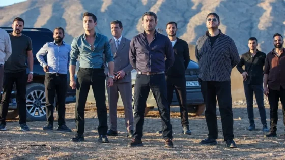 Group of men standing in a barren desert with rocky hills behind, some in dress shirts and suits, facing the camera with cars in the background