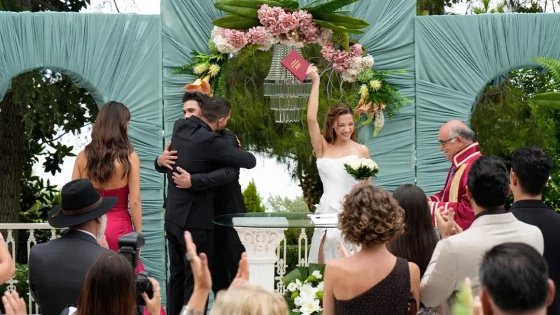 Bride in a white off-shoulder gown raises a pink book above her head under a teal fabric arch as guests applaud at an outdoor wedding.