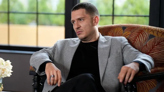 Man in a light gray blazer and black shirt sits in a patterned armchair with a serious, contemplative expression by a windowed room.