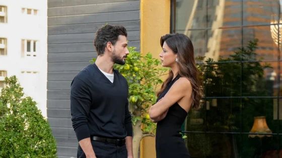 Man and woman stand face-to-face outdoors, talking seriously beside a gray wall with greenery nearby.