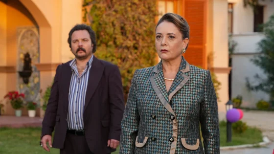 Woman in a gray patterned coat with pearls, looking serious, in the foreground; a man in a dark suit stands blurred behind her in a sunny courtyard.